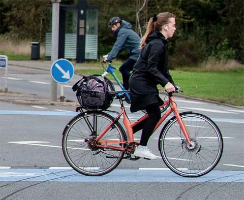 Foto af en ung cyklist, der cykler over krydset Stormgade/Spangsberg Møllevej med airpods i ørerne og en taske i cykelkurven. 